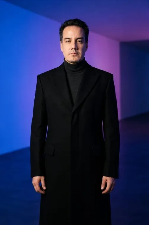 Edson Noyola de cuerpo entero con abrigo largo, sobre fondo azul y violeta